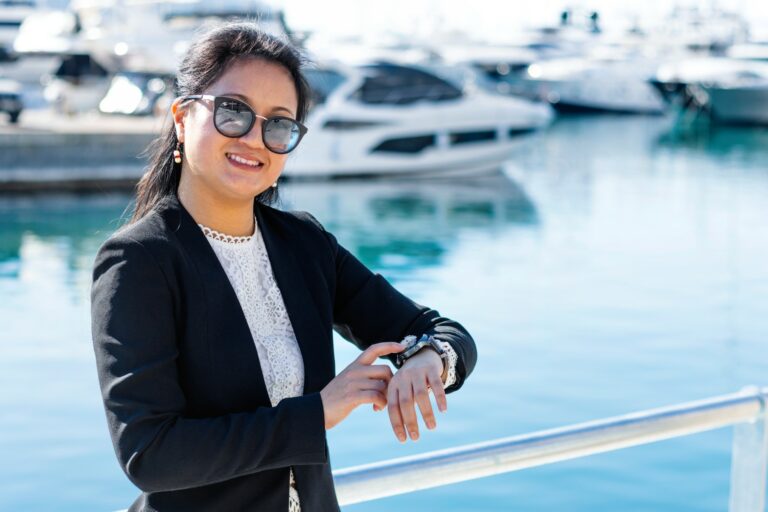 A woman in business attire pointing at her watch while standing on a boat with luxury boats in the background.